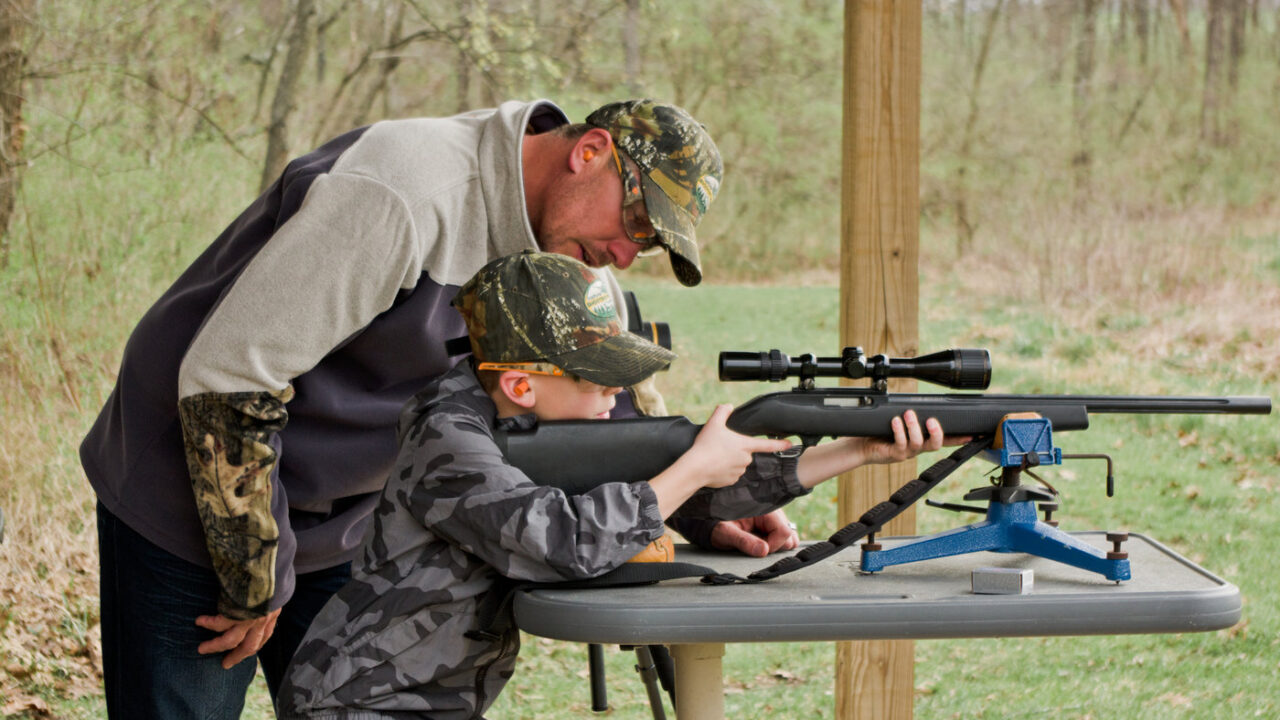 An adult trains a youth on how to safely shoot a rifle at a shooting range.
