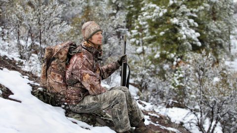 gun hunter wearing camouflage and sitting in snowy scene holding a muzzleloader rifle